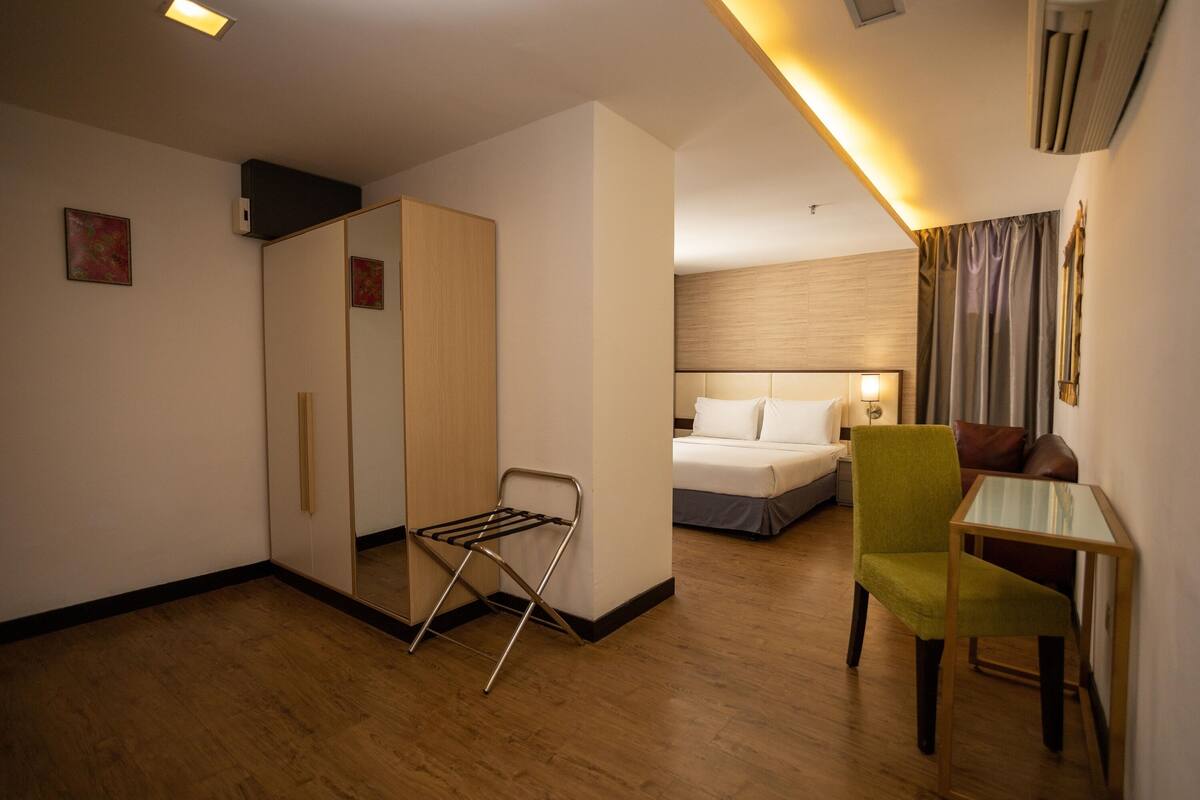 A hotel room with a bed, a green chair, a small table, a mirror, and a wardrobe.