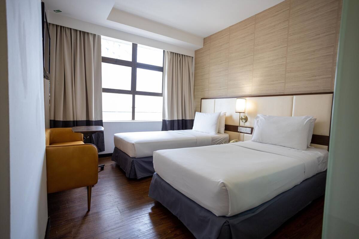 A hotel room with two beds, a small table, a chair, and a large window with curtains.