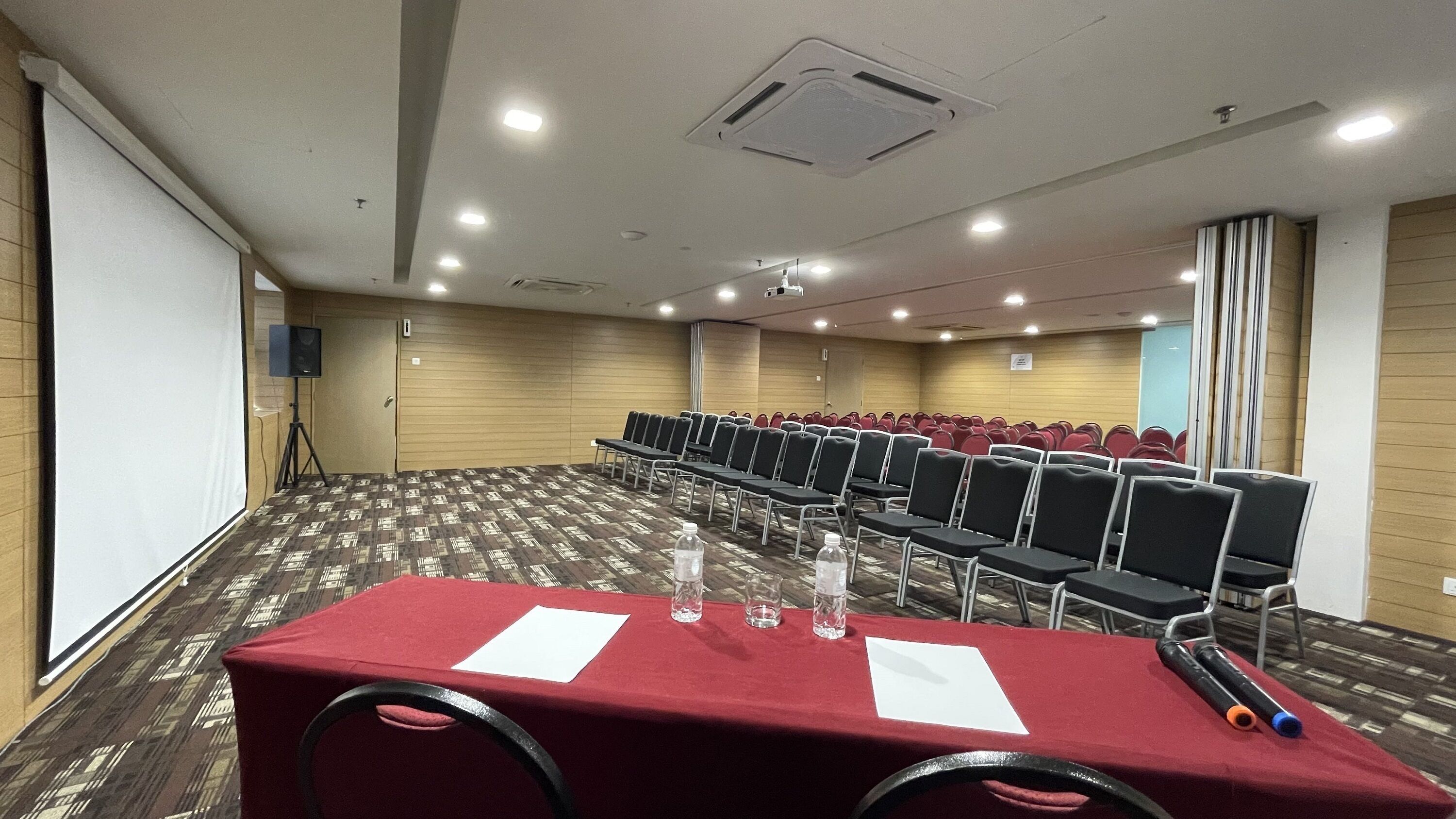 Una sala de conferencias con filas de sillas, una mesa con papeles y botellas de agua, y una pantalla para proyector.