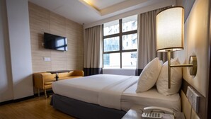 A hotel room with a bed, a bedside table with a phone, a TV mounted on the wall, and a window with curtains.