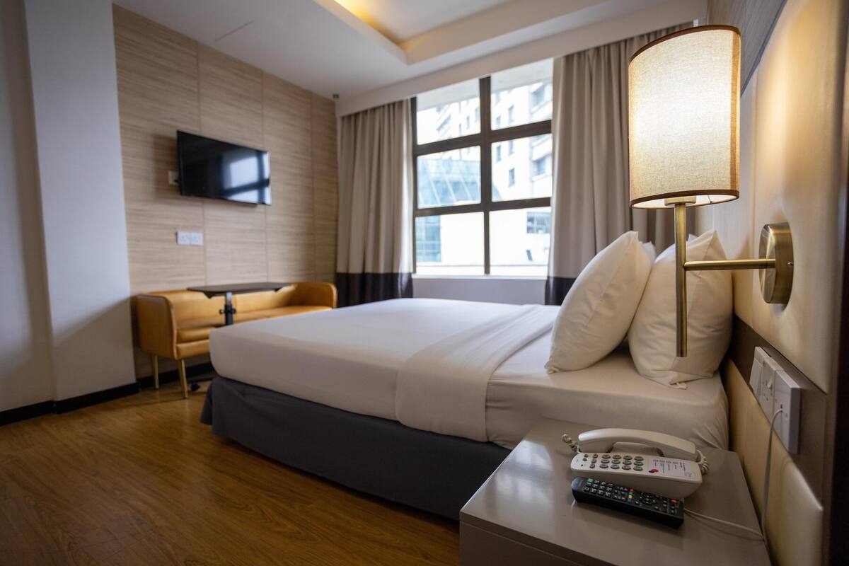 A hotel room with a bed, a bedside table with a phone, a TV mounted on the wall, and a window with curtains.