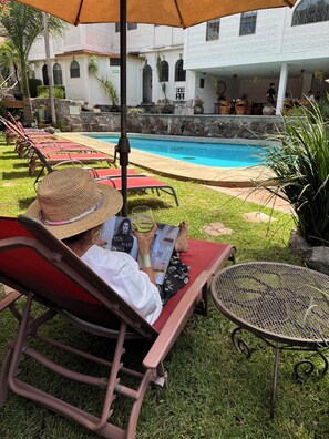 A person relaxing on a red lounge chair with a straw hat, reading a magazine. A pool is visible in the background, surrounded by a white building with arched windows. - Hotel Ilebal Cuernavaca Centro - un lugar con encanto (Cuernavaca)