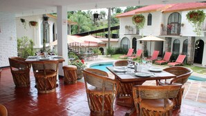 An outdoor dining area with wicker furniture, a pool, and a view of a white building with red roofing.