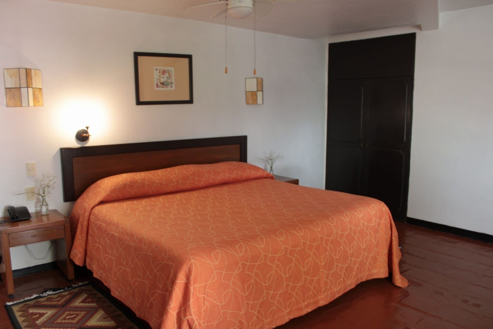 A neatly made bed with an orange patterned bedspread, a wooden nightstand with a lamp, a framed picture on the wall, and a door to the right.