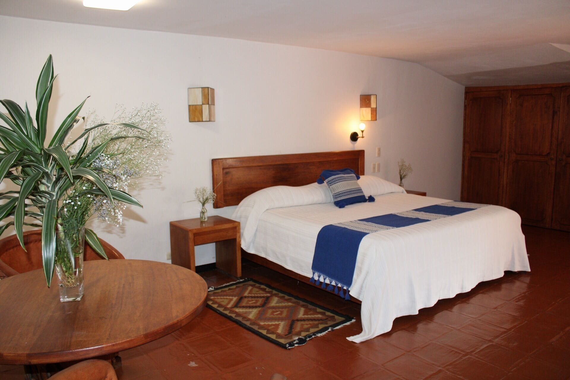 Een netjes opgemaakte slaapkamer met een houten hoofdbord, een bed met wit beddengoed en blauwe accenten, een nachtkastje met een lamp en een kamerplant.