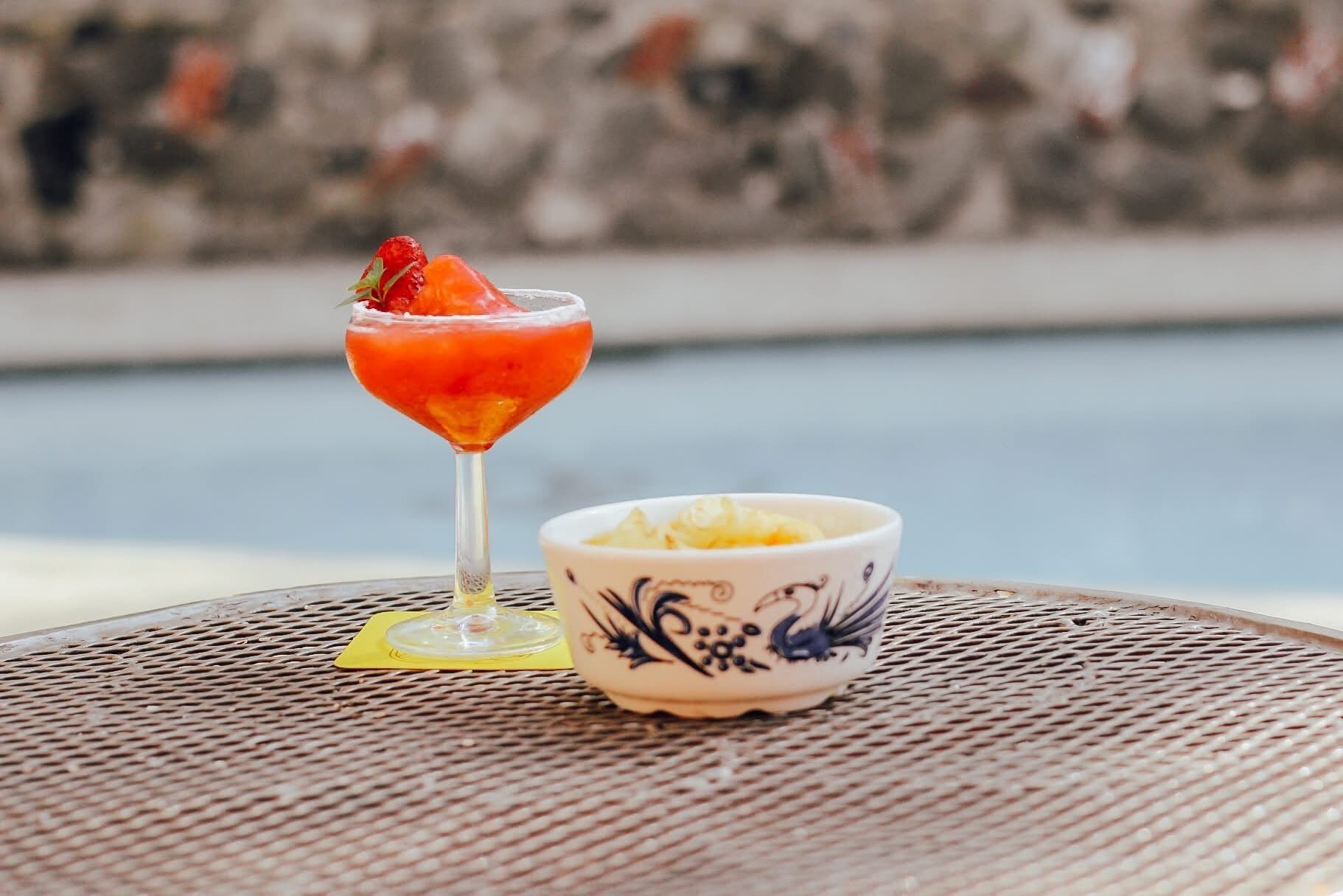 A cocktail glass with a strawberry garnish on a metal table next to a decorative bowl.