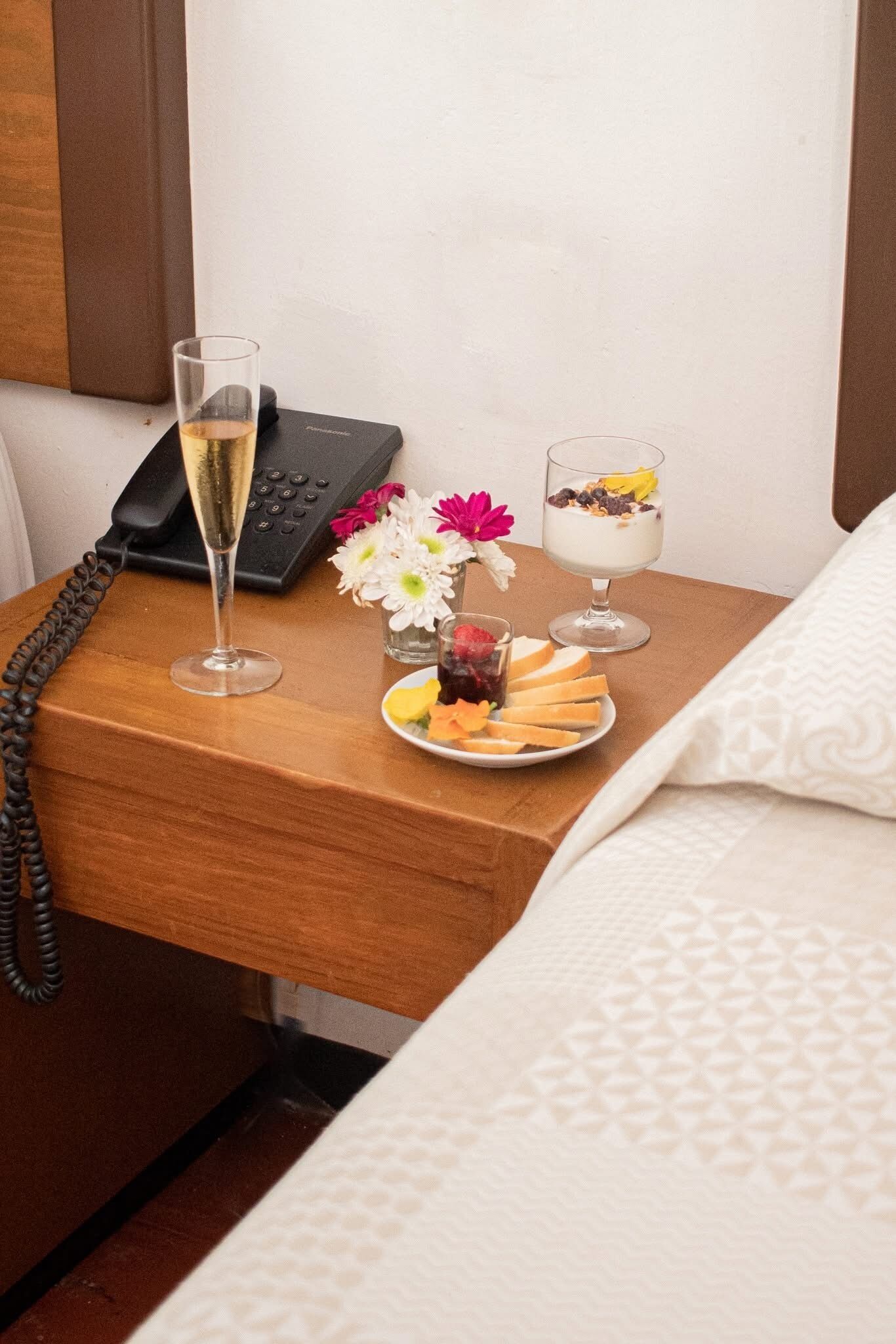 A neatly arranged hotel room with a wooden nightstand holding a phone, a glass of champagne, a small vase with flowers, and a plate of fruit and pastries.