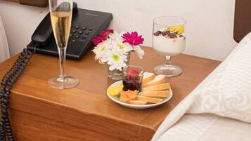 A neatly arranged hotel room with a wooden nightstand holding a phone, a glass of champagne, a small vase with flowers, and a plate of fruit and pastries.
