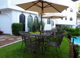 Un patio con una mesa y sillas, rodeado de follaje y un edificio blanco con ventanas arqueadas.