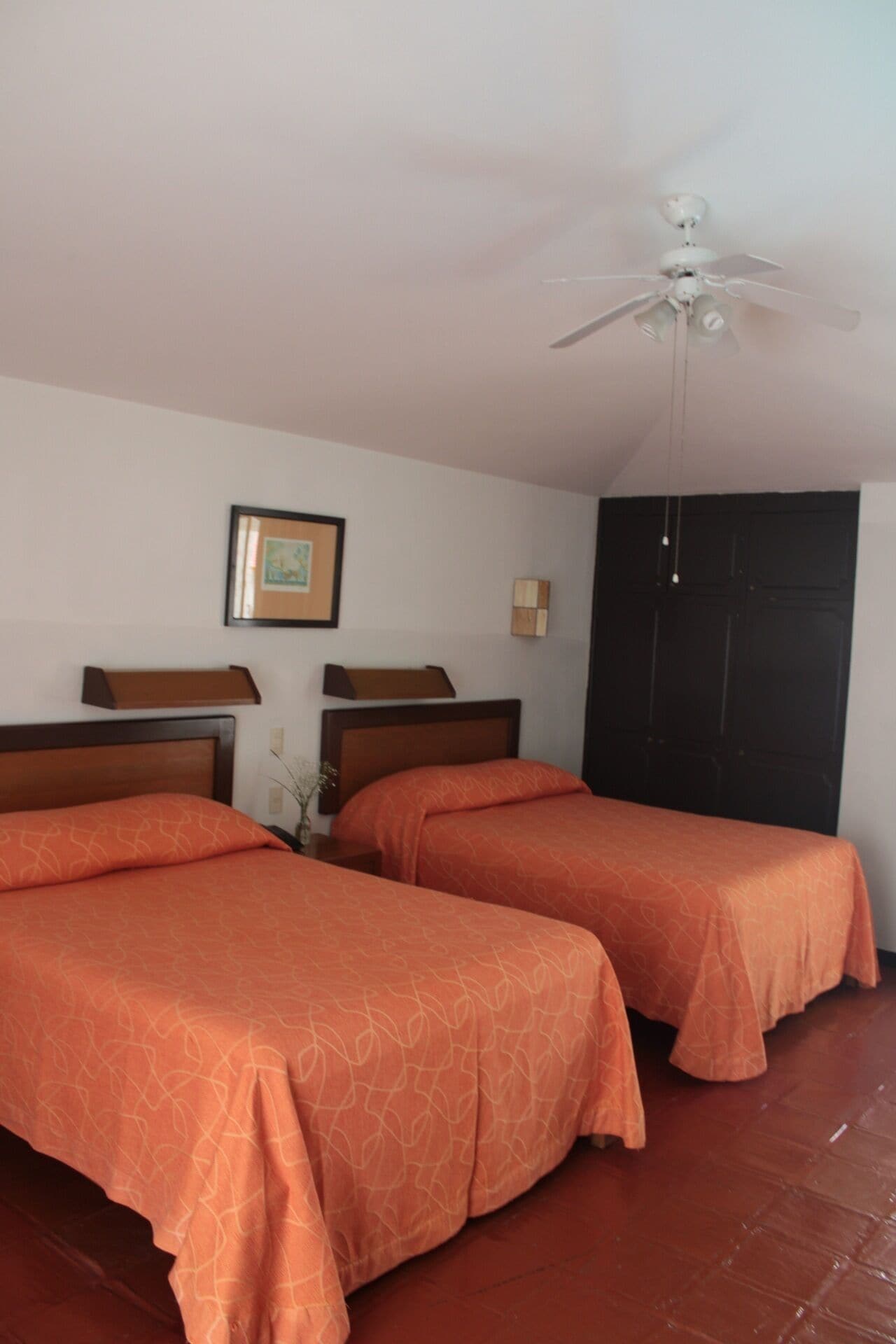 Quarto de hotel com duas camas, um ventilador de teto, um quadro na parede e uma porta escura.