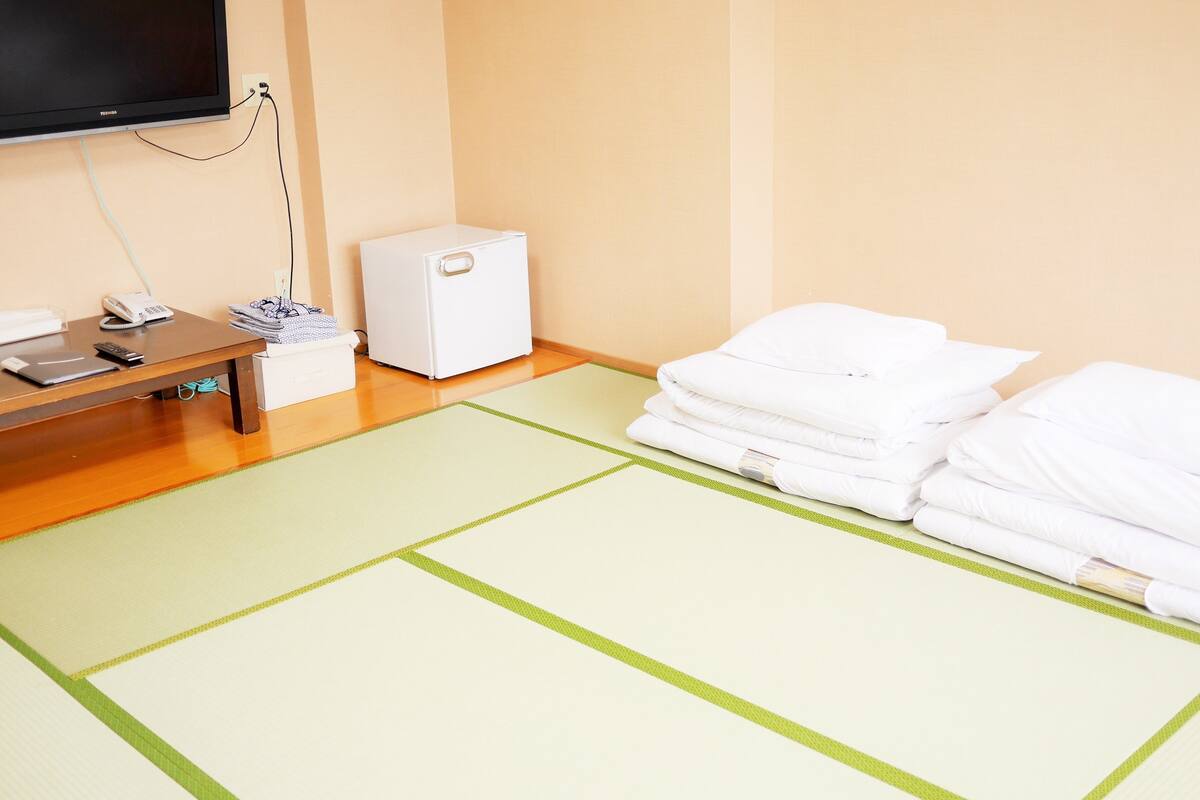 Habitación de hotel con televisor de pantalla plana, un pequeño electrodoméstico blanco, teléfono y toallas dobladas con cuidado sobre un esterilla de tatami.