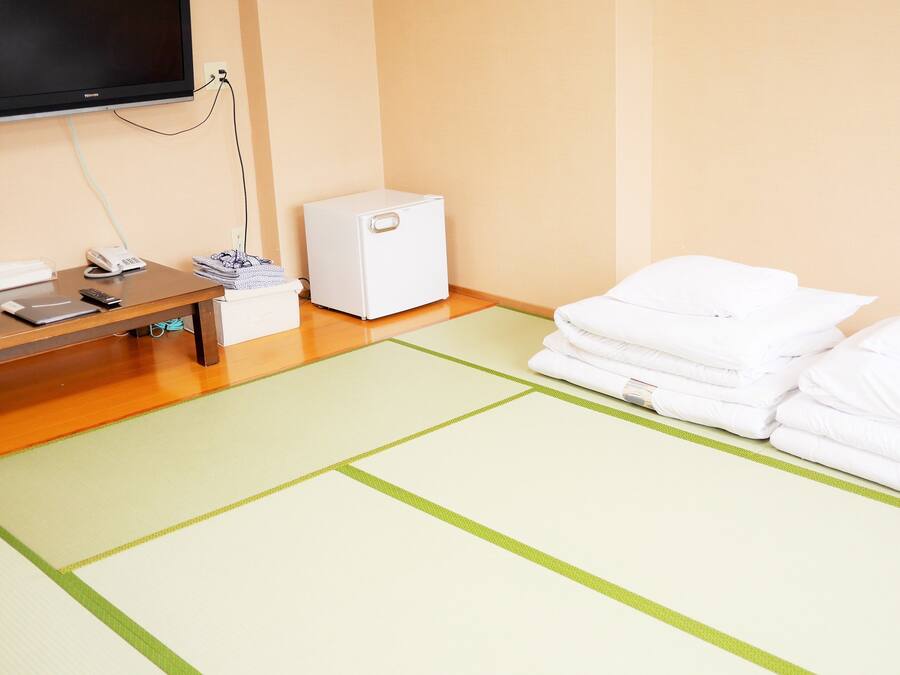 A hotel room with a flat-screen TV, a small white appliance, a telephone, and neatly folded towels on a tatami mat.