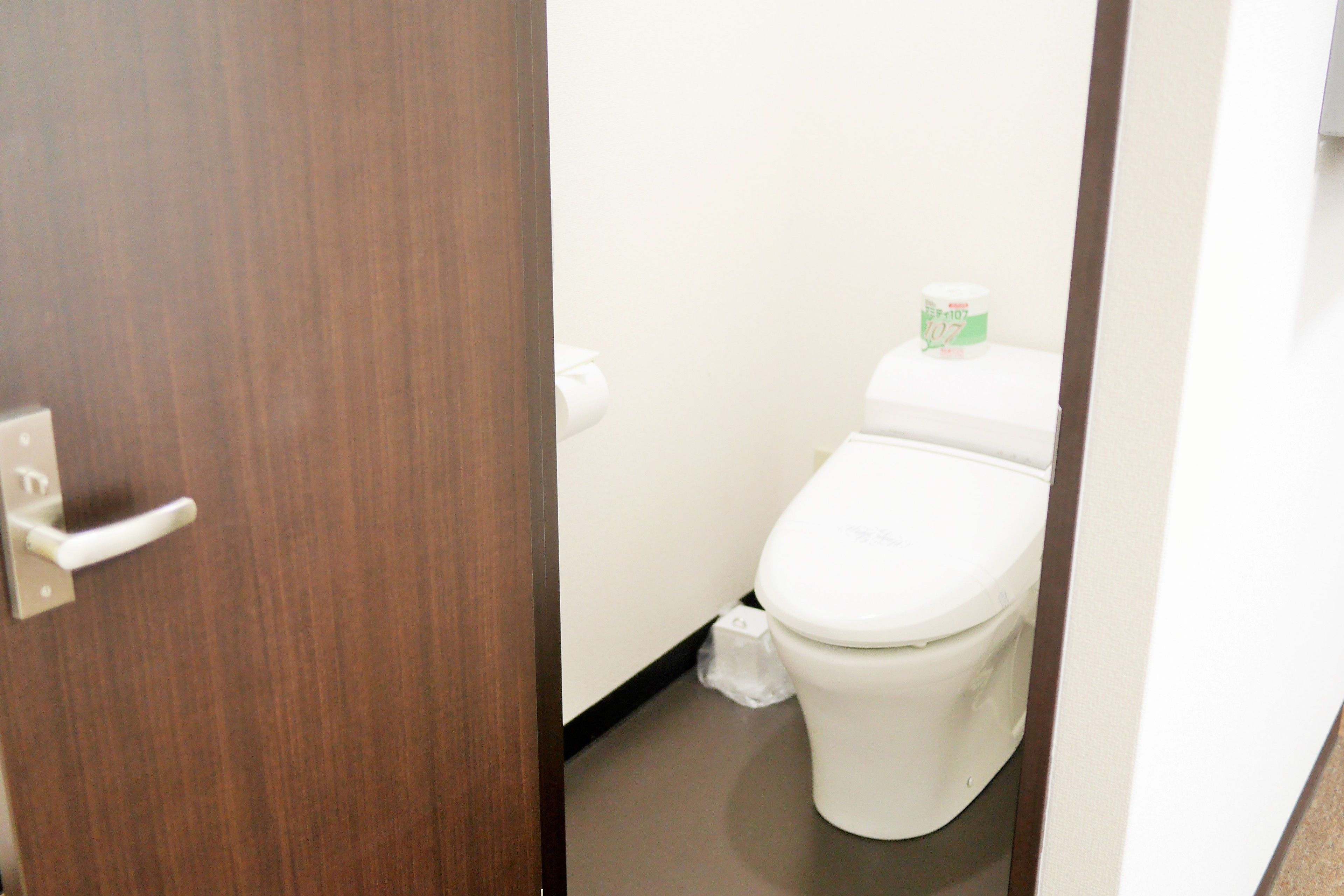Un baño pequeño y privado con inodoro, un dispensador de papel higiénico blanco y un pequeño cubo de basura.