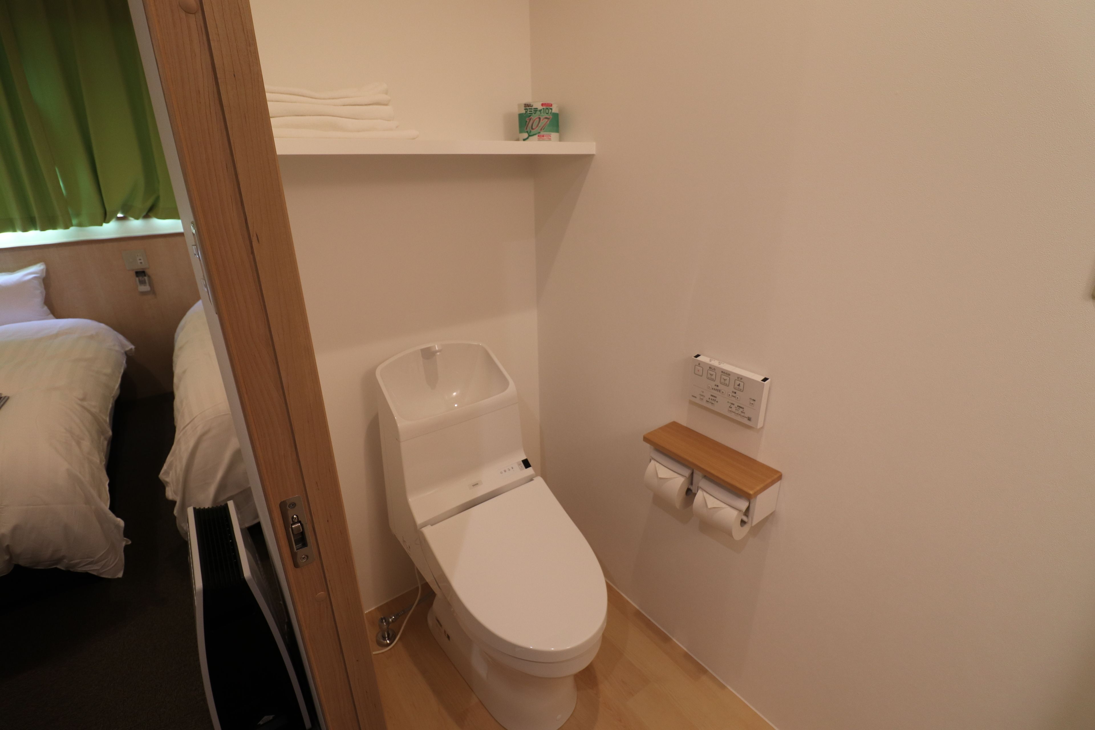 A small, clean bathroom with a toilet, a shelf with toiletries, and a bed in the adjacent room.