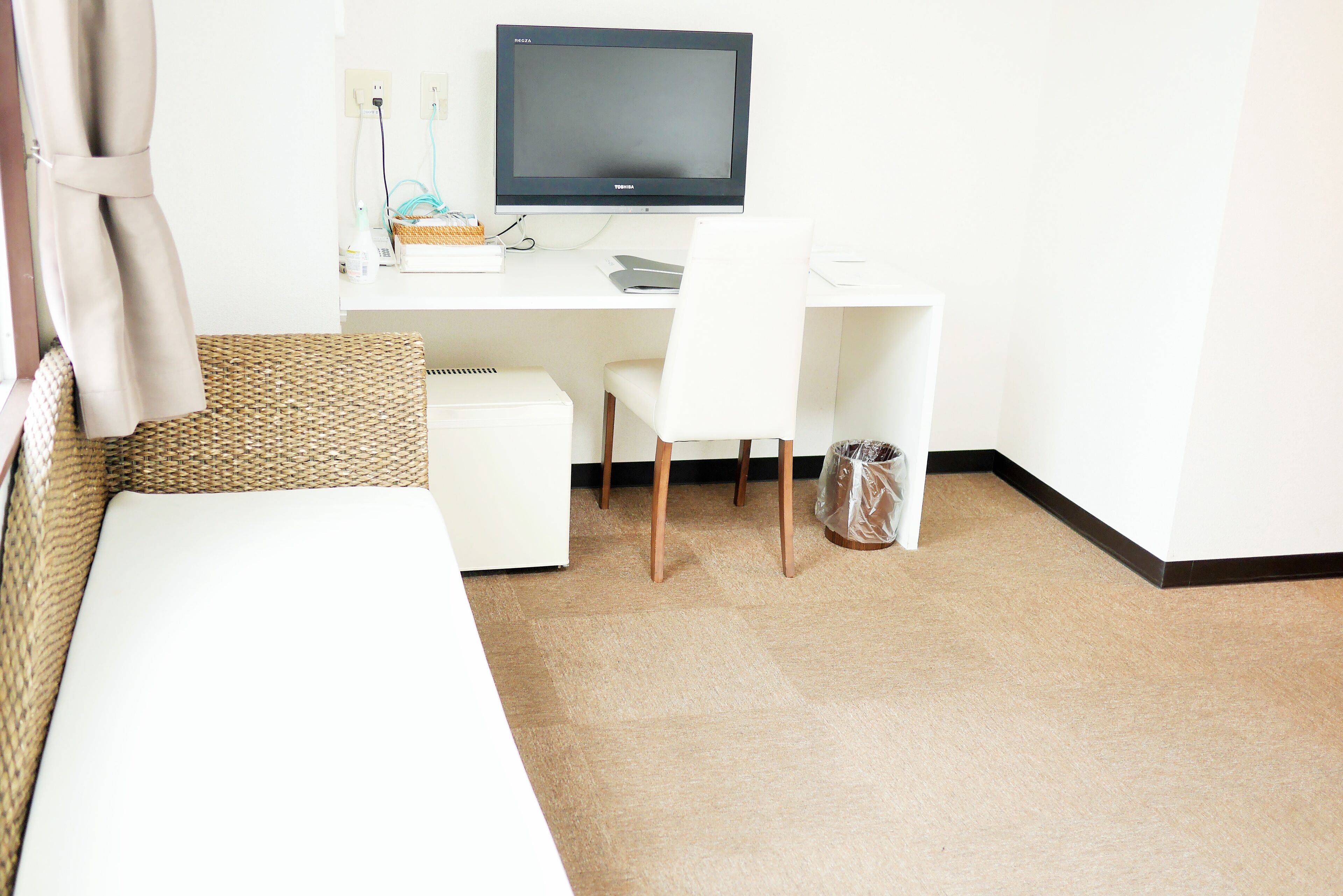Une chambre d’hôtel avec un bureau blanc, un ordinateur, une chaise blanche, une petite poubelle et un lit blanc.