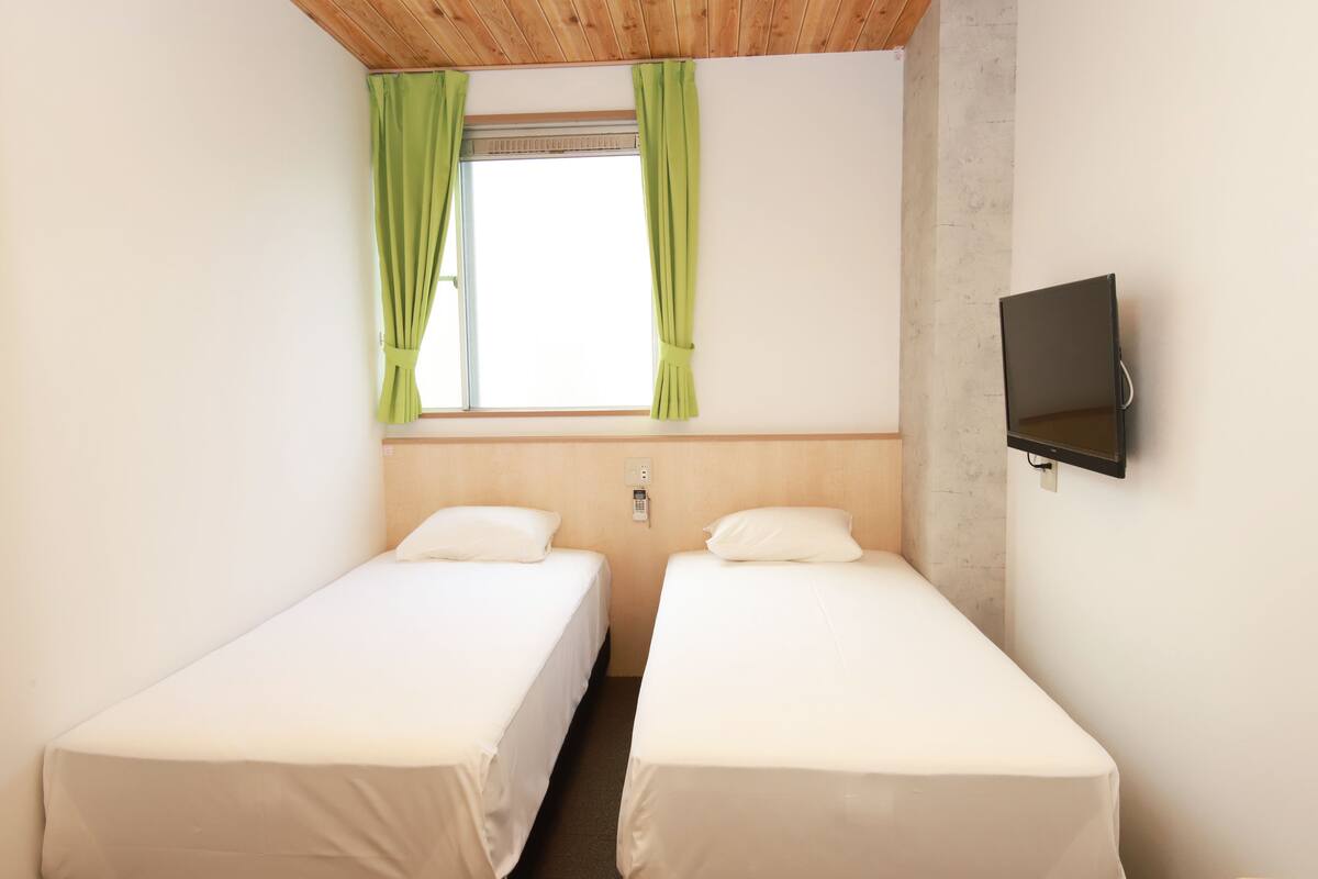 Una habitación con dos camas individuales, una ventana con cortinas verdes, un televisor de pared y techo de madera.