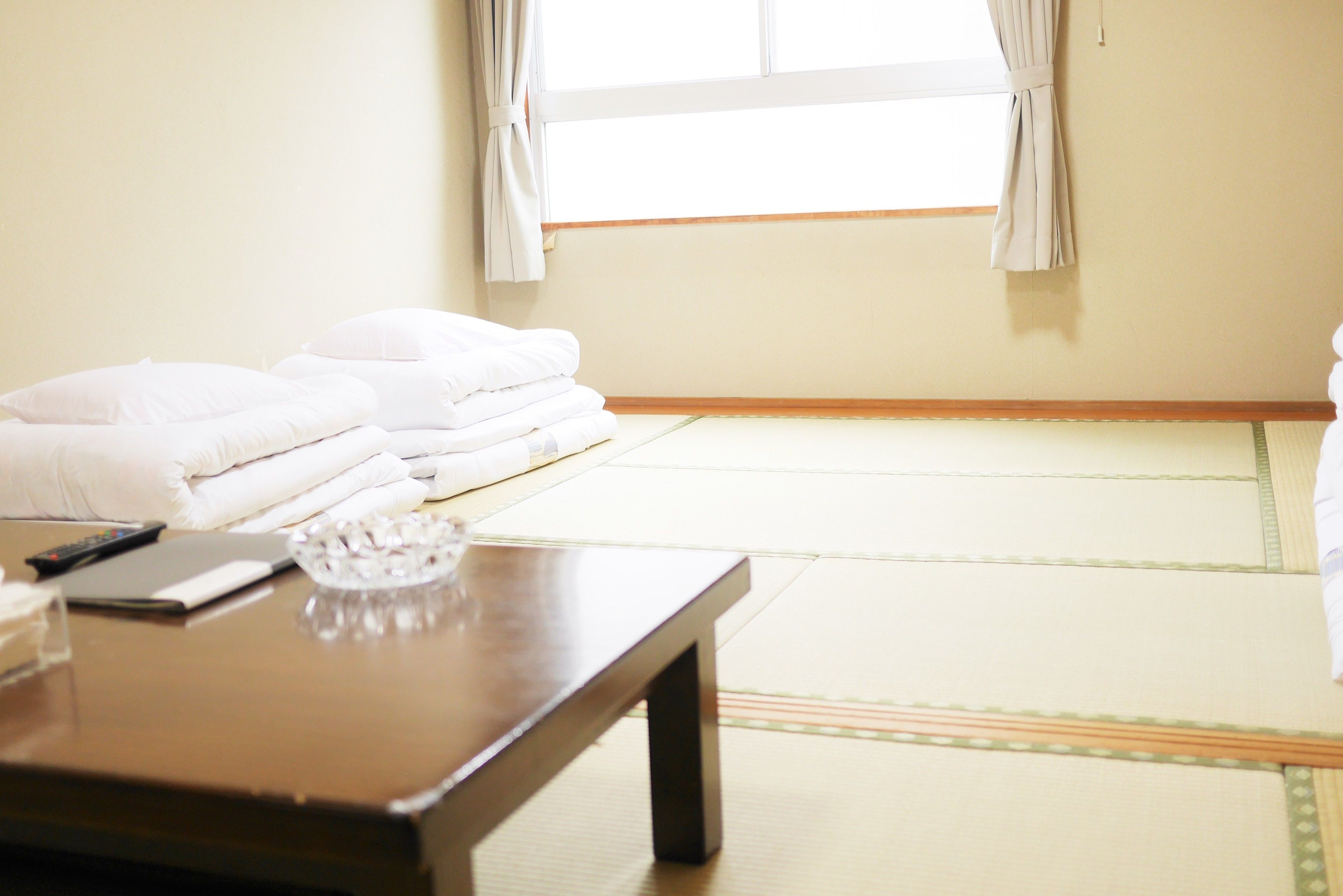 Una habitación con esteras de tatami, una cama con ropa de cama blanca, una mesita con un control remoto, un vaso y un bol.