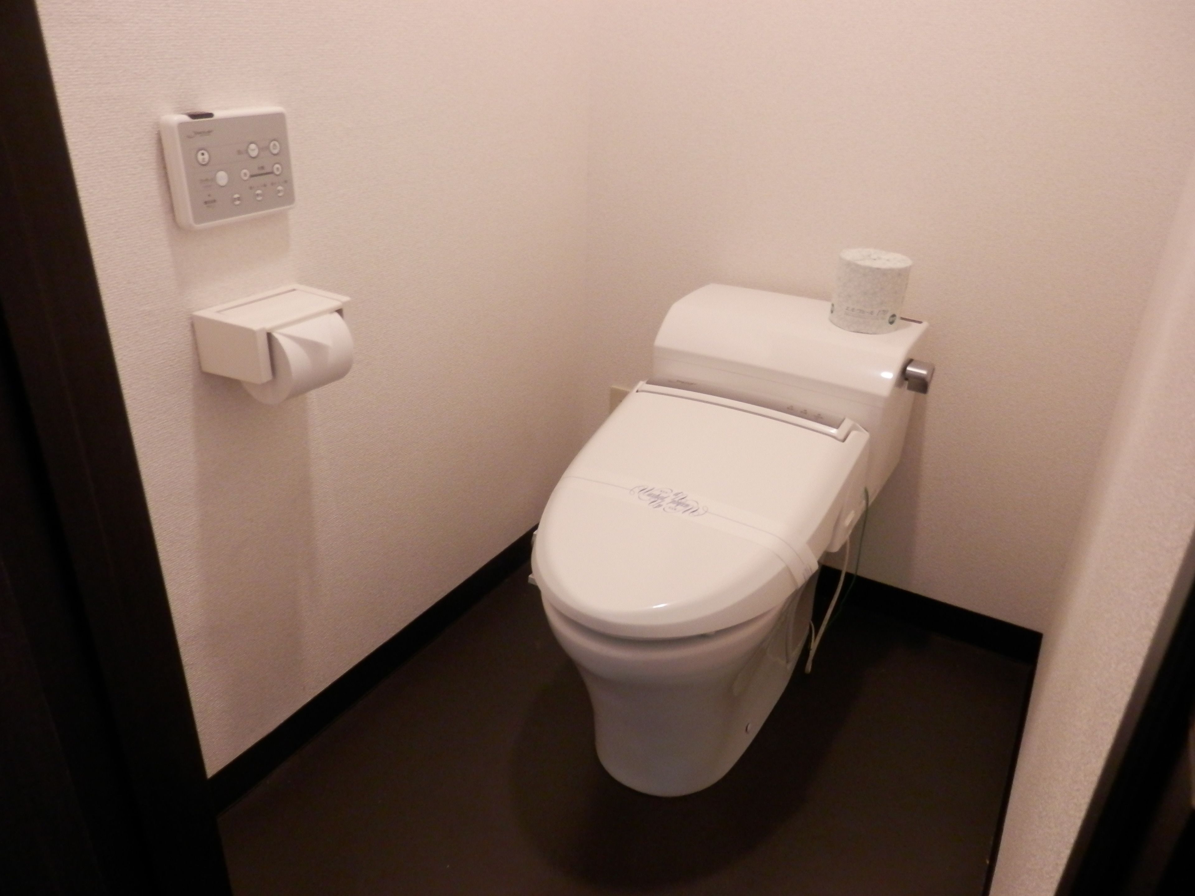 Inodoro blanco estándar con palanca de descarga y un dispensador de toallas de papel en la pared.
