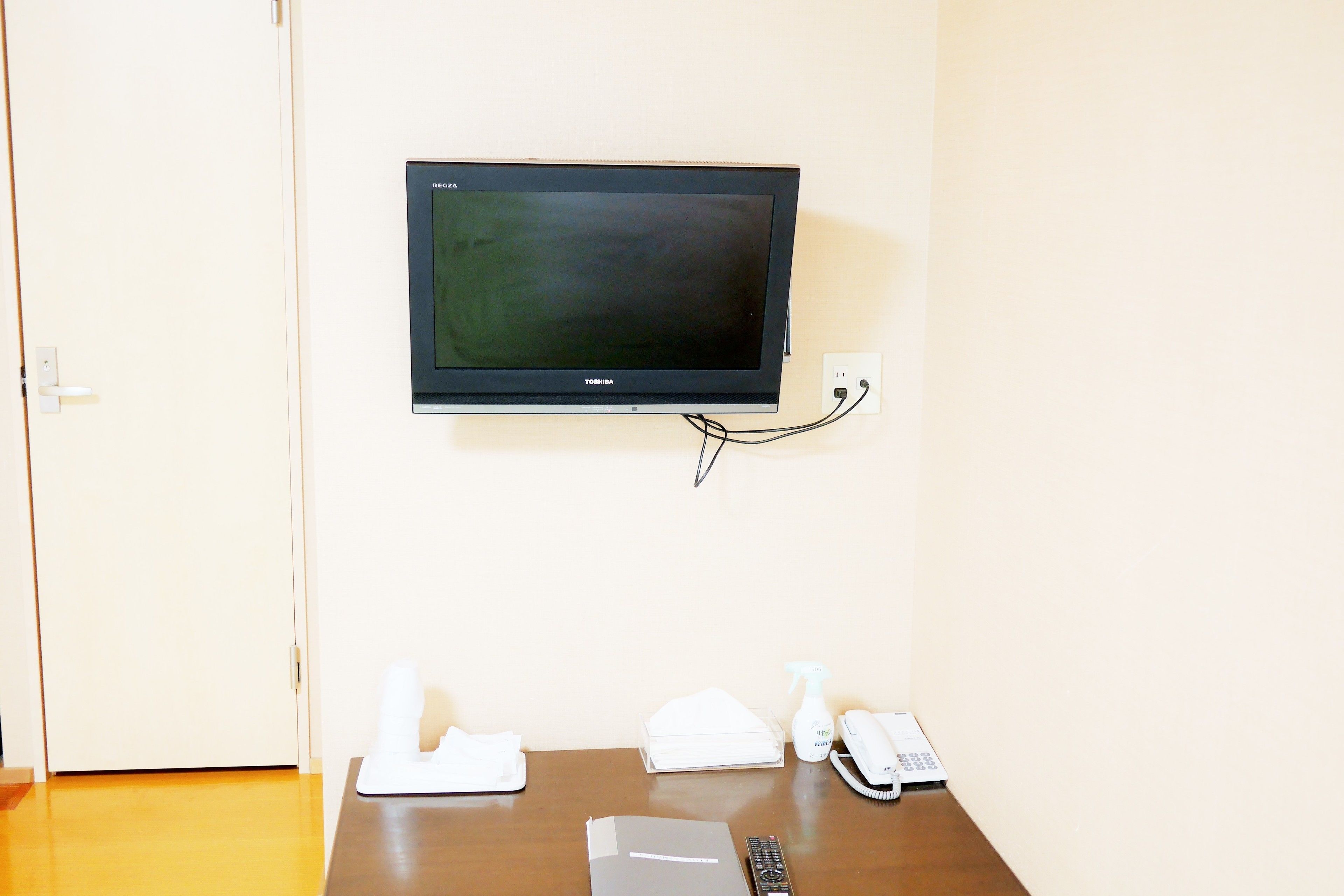 A room with a mounted television on the wall, a wooden table with a remote control, a telephone, and a spray bottle.