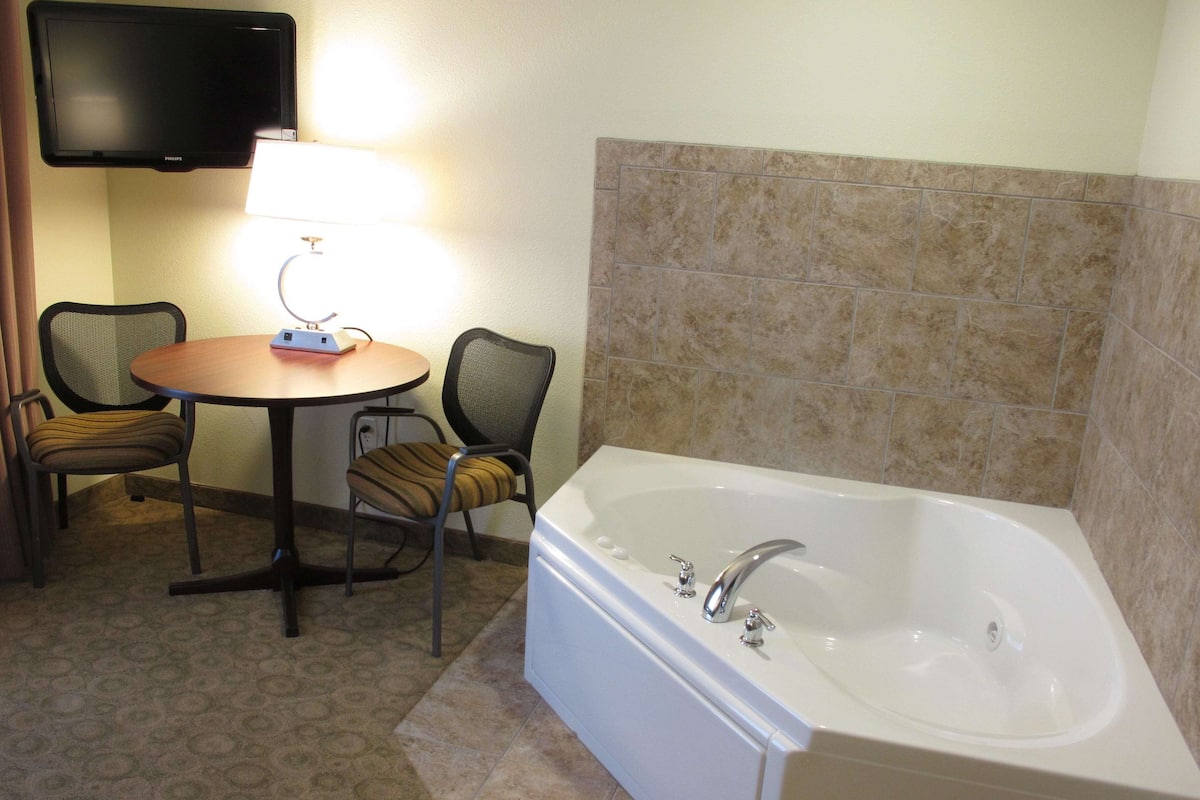 Une salle de bain avec une baignoire blanche, deux chaises, une petite table avec une lampe et un téléviseur à écran plat fixé au mur.