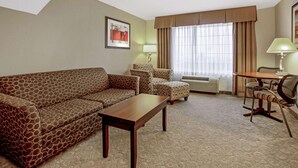 A hotel room with a sofa, armchair, ottoman, a small table, a desk with a chair, a lamp, and a large window with curtains. - La Quinta Inn & Suites by Wyndham Bismarck (Bismarck)