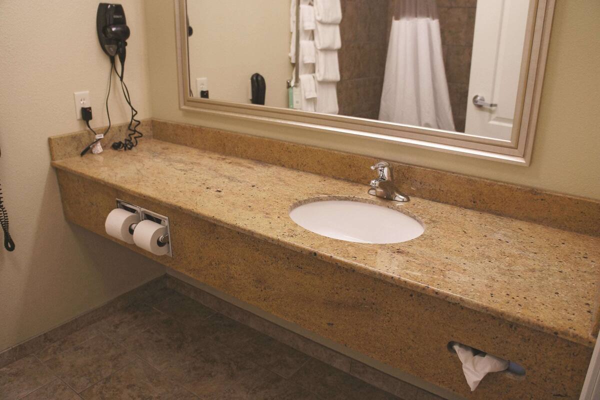 Une salle de bain dotée d’un plan de toilette en granit, d’un lavabo, d’un miroir, d’un sèche-cheveux et d’un porte-serviettes.