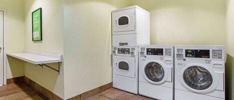 A laundry room with stacked washers and dryers, a white shelf, and a green sign on the wall.