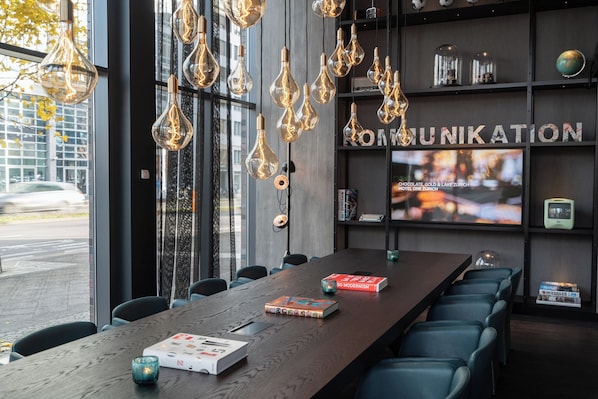 A modern conference room with a long table, blue chairs, hanging light fixtures, and a bookshelf with books and a globe. - Motel One Berlin-Spittelmarkt (Berlin)
