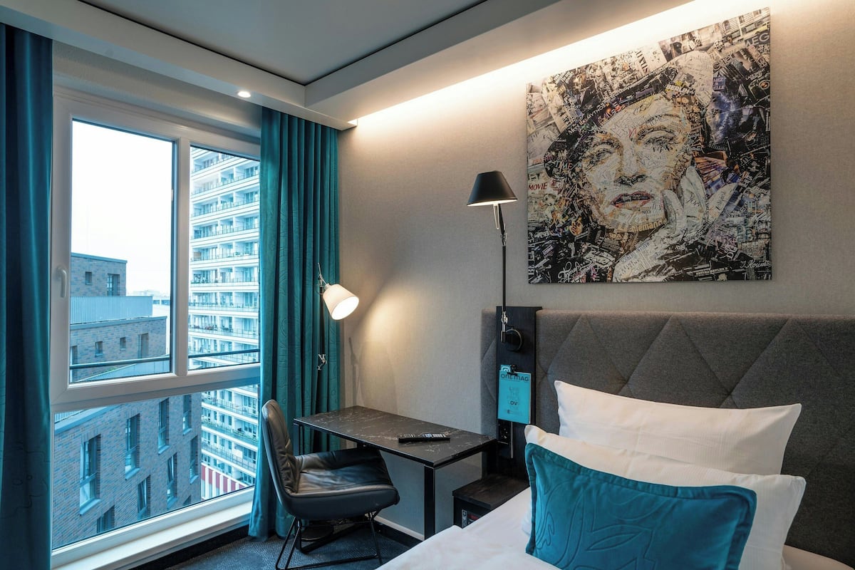 A hotel room with a bed, a desk, a chair, a window with curtains, a wall-mounted lamp, and a large abstract painting.