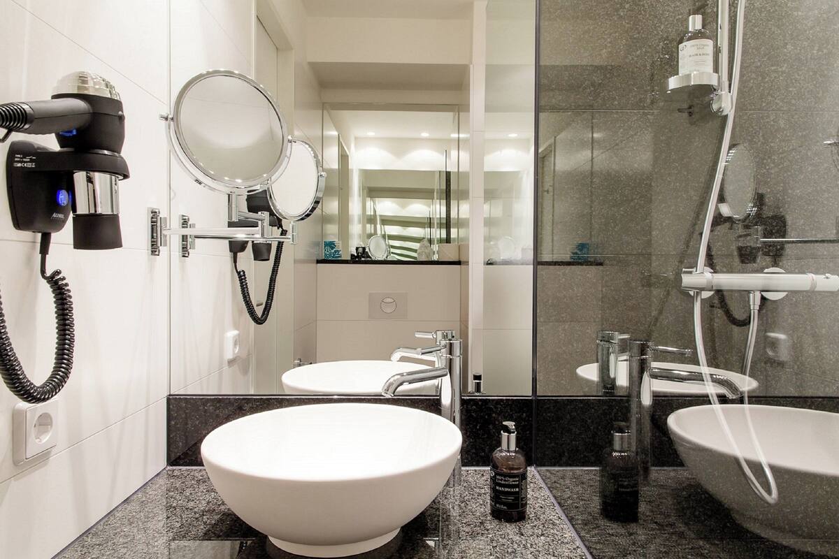 A modern bathroom with a large white sink, two mirrors, a hairdryer, and a shower area.