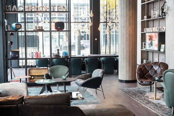 A modern lounge area with leather sofas, a coffee table, and a bookshelf filled with books and decorative items. - Motel One Berlin-Spittelmarkt (Berlin)