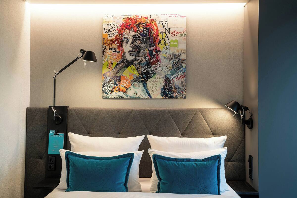 A hotel room with a tufted headboard, a bed with white and teal pillows, a nightstand with a lamp, and a wall art piece above the bed.