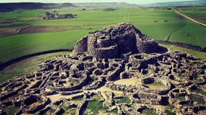 Vue aérienne d’une ancienne ruine en pierre, avec des structures circulaires et un monticule central, le tout entouré de champs verdoyants et d’une route.