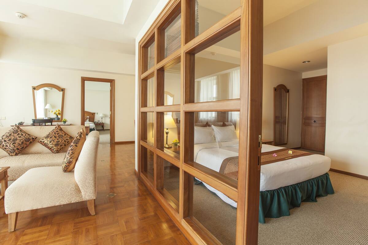 Een hotelkamer met een raam met houten kozijn, een bank, een bed met groene laken en een spiegel.