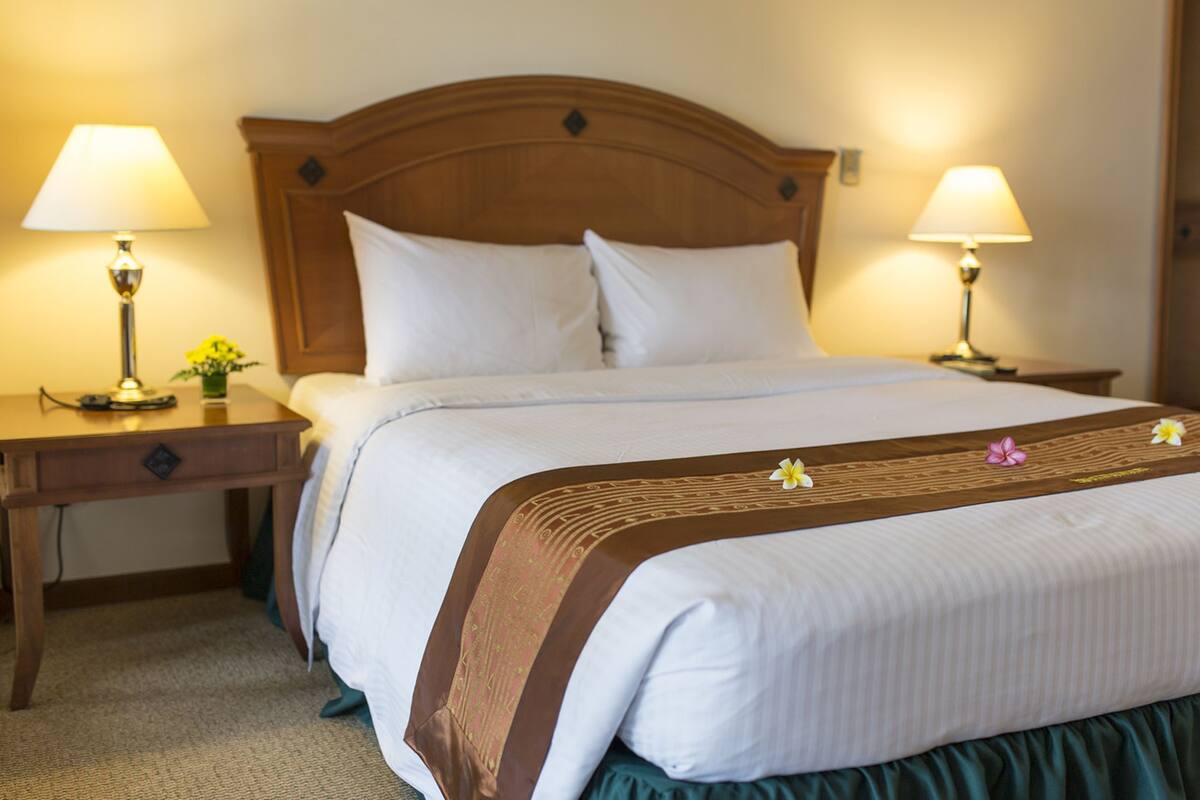 Een hotelkamer met een houten hoofdeinde, twee bedlampen, een bed met witte lakens en een decoratief kussen.