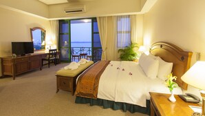 A hotel room with a bed, a desk with a TV, a chair, a bedside table with a lamp, and a view of the sea.