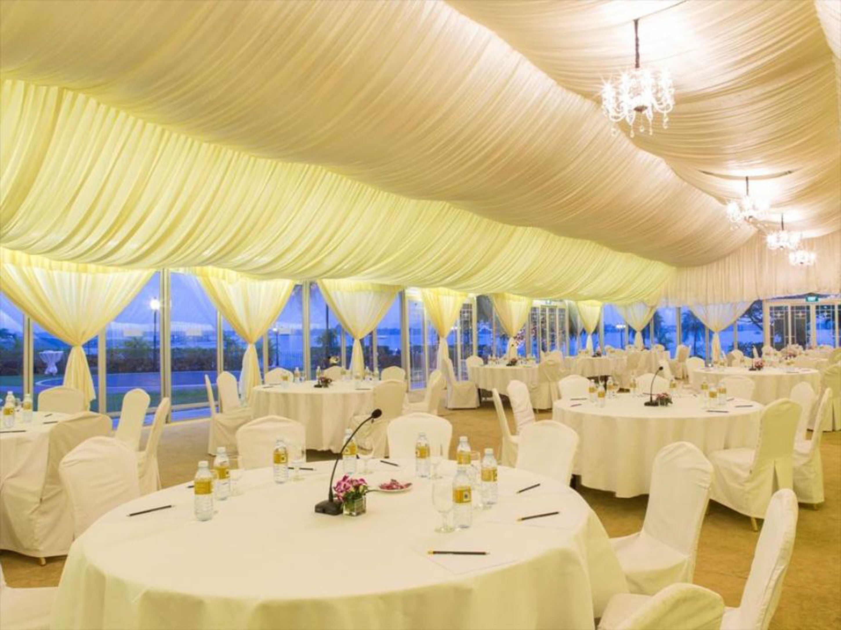 Un espacio para eventos con carpas, mesas redondas, sillas blancas y candelabros.