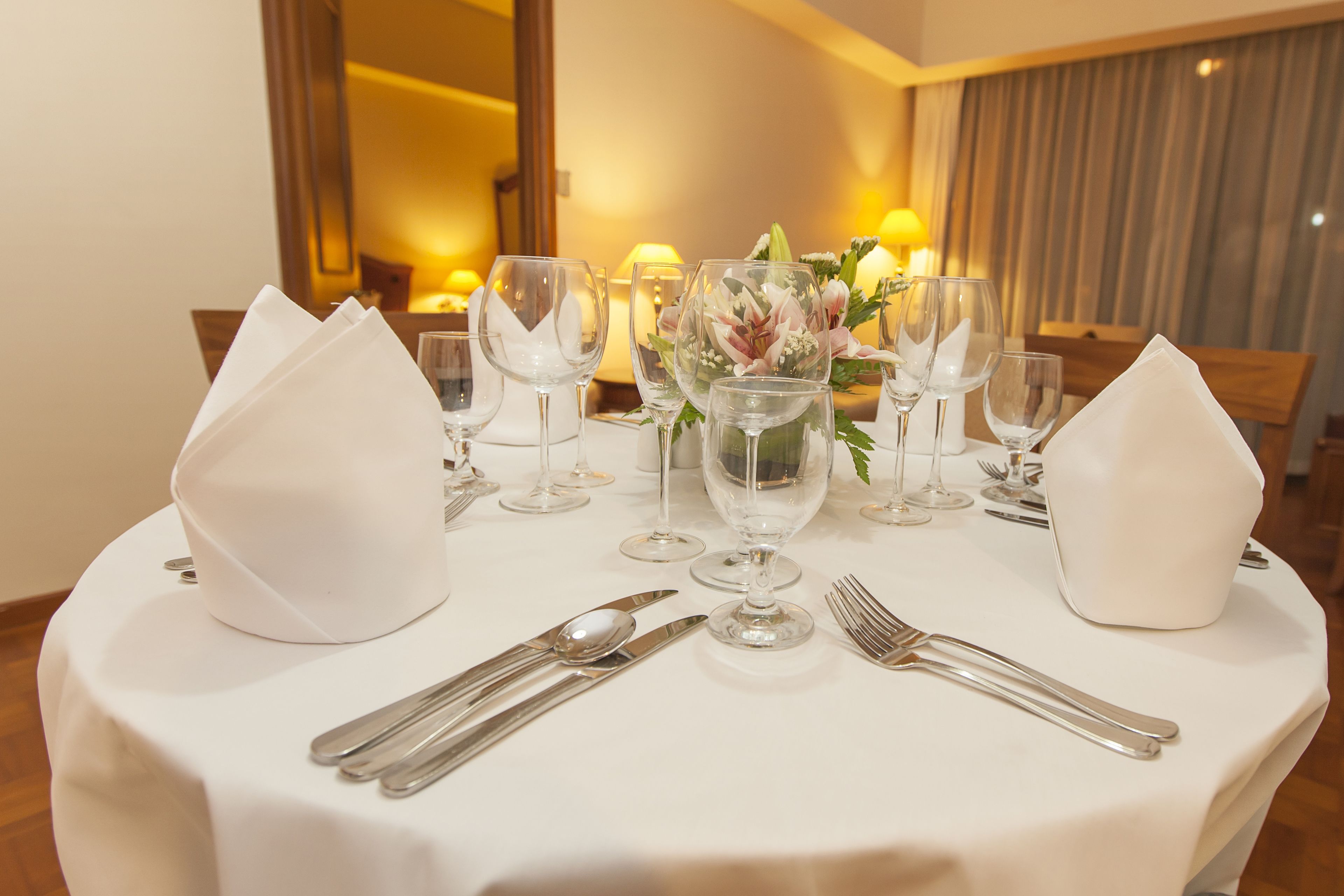 A round dining table set with white napkins, silverware, and wine glasses, centered with a floral arrangement.