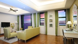 A hotel room with a sofa, armchairs, a dining table, a TV, and a view of the sunset through large windows.