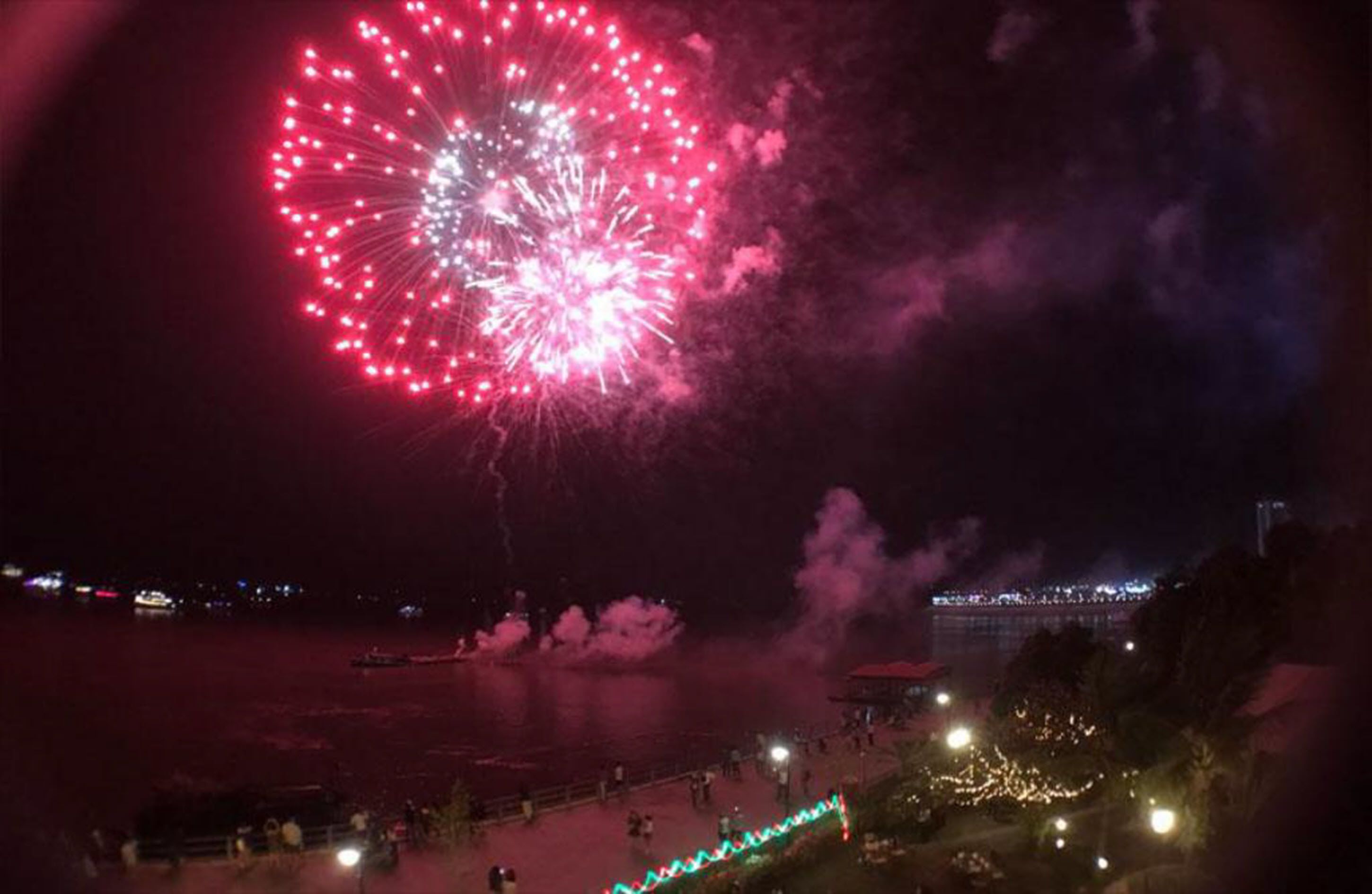 A nighttime view of a waterfront with fireworks display, a dock, and a promenade with lights.