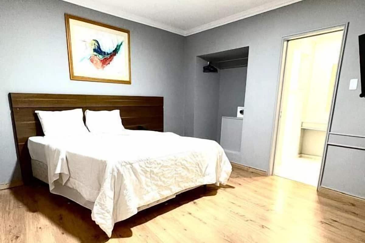 Une chambre avec un plancher en bois, un lit recouvert de draps blancs, un tableau encadré au mur et une porte sur le côté droit.