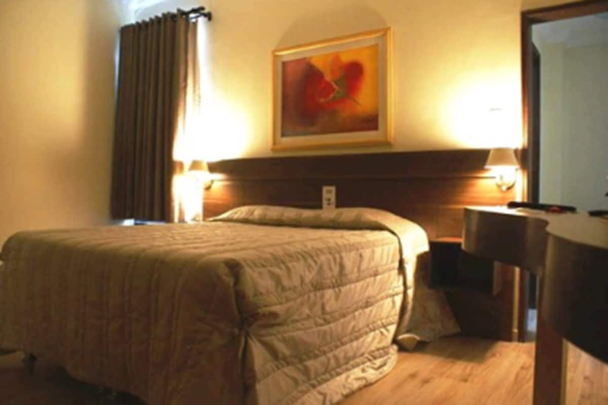 Une chambre d’hôtel avec un lit, une table de chevet, une lampe, un tableau au mur et une fenêtre avec des rideaux.