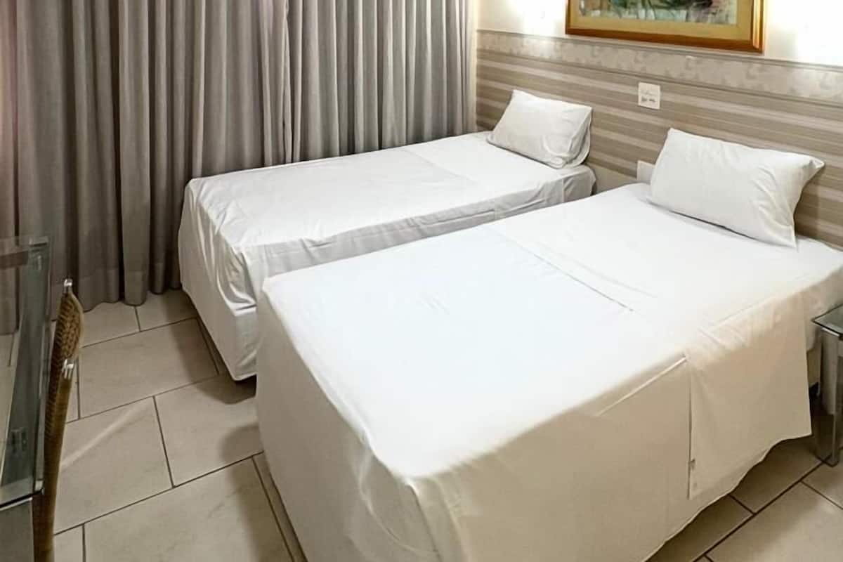 Une chambre d’hôtel avec deux lits simples, une grande fenêtre avec des rideaux, un tableau au mur et une petite table avec une chaise.