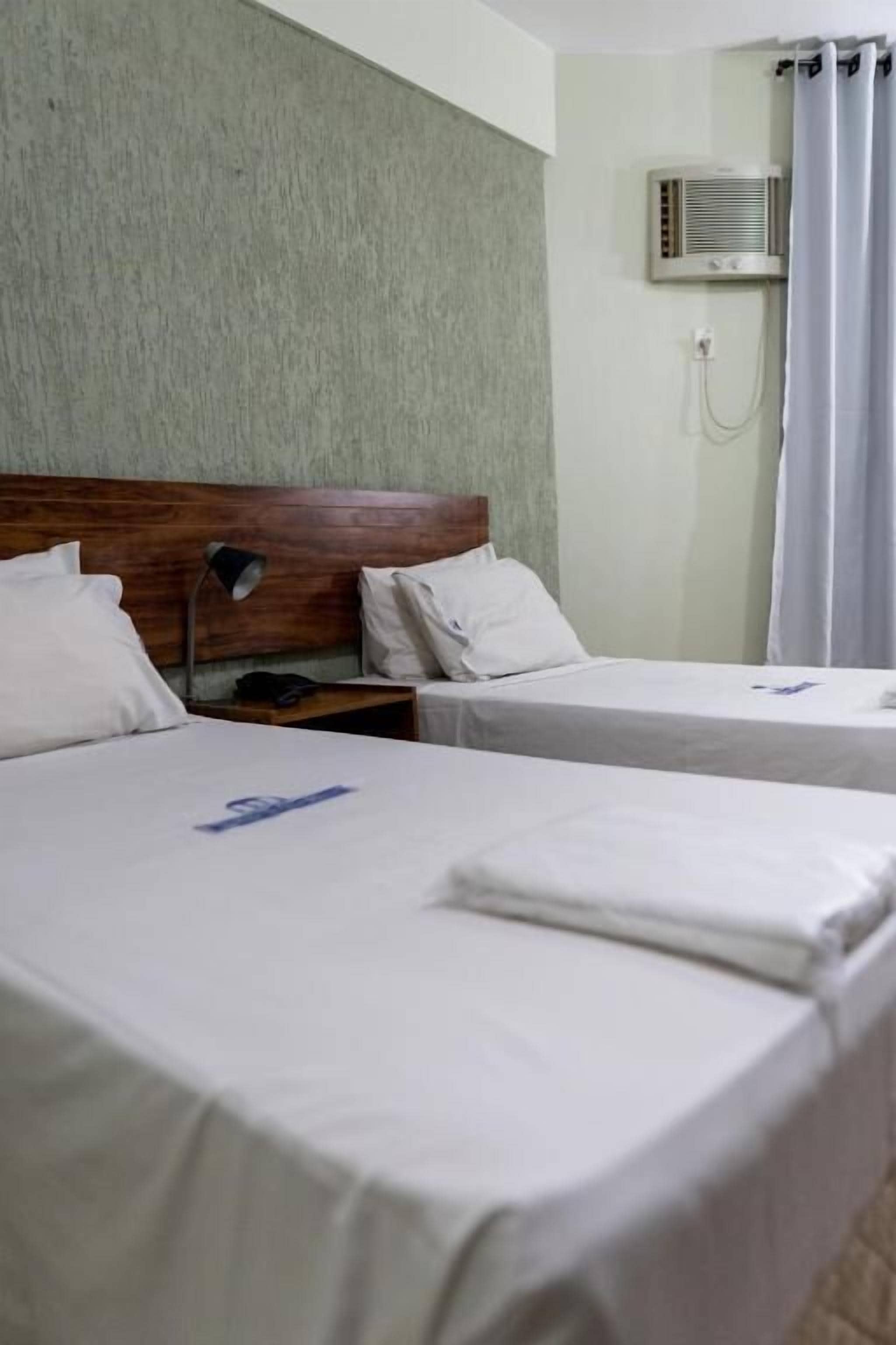 A hotel room with two beds, a wooden headboard, a nightstand, a lamp, an air conditioning unit, and a curtain.