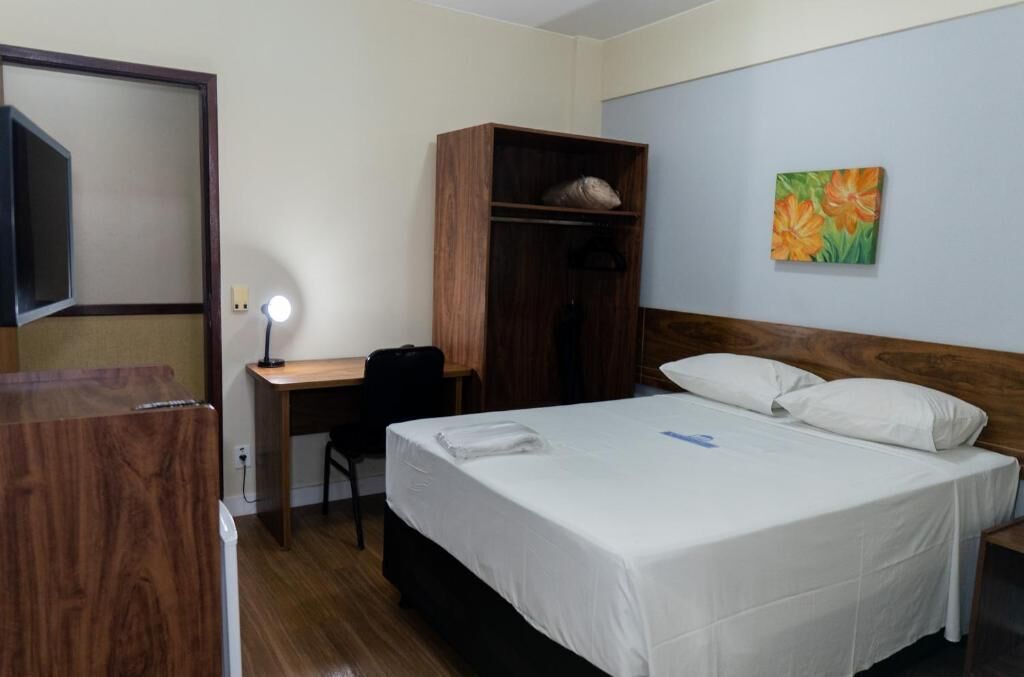 A hotel room with a bed, a desk, a wardrobe, and a painting on the wall.