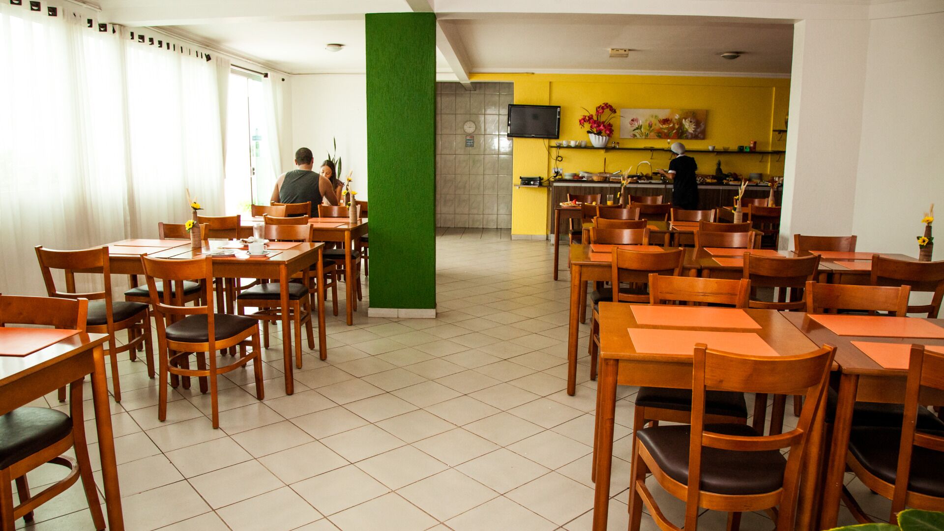 Un restaurant amb taules i cadires de fusta, una zona de cuina i un pilar verd.