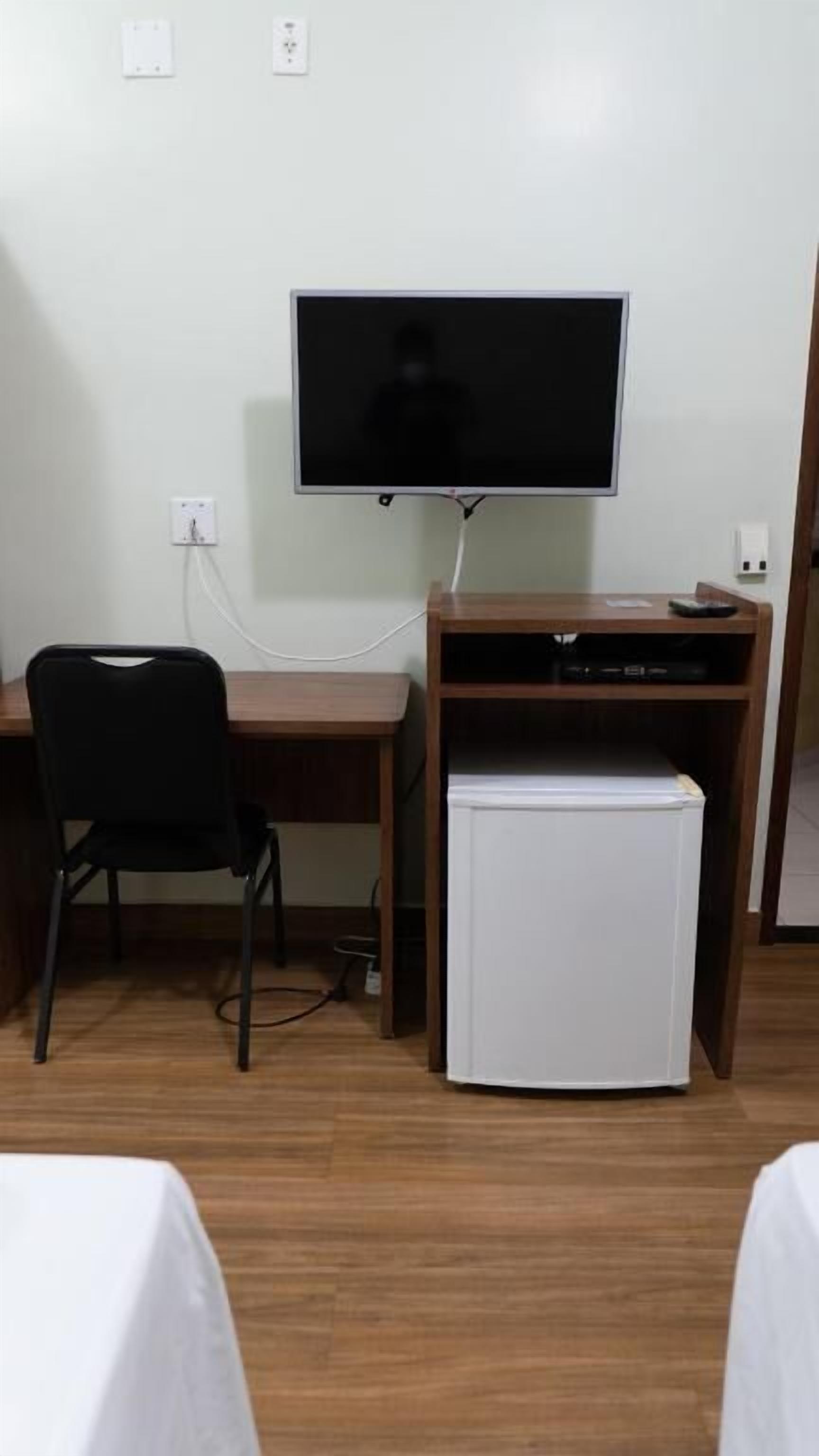 A room with a wooden desk, a black chair, a television mounted on the wall, and a small white refrigerator.