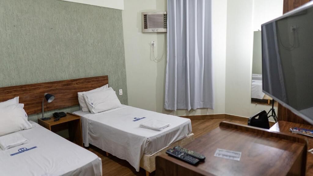 A hotel room with two single beds, a wooden headboard, a desk with a computer and a TV, an air conditioning unit, and a curtain.