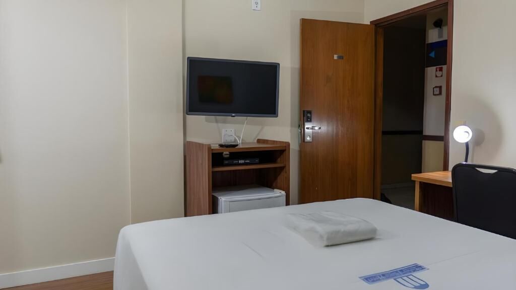 A hotel room with a bed, a television mounted on the wall, a wooden wardrobe, a desk with a chair, and a desk lamp.