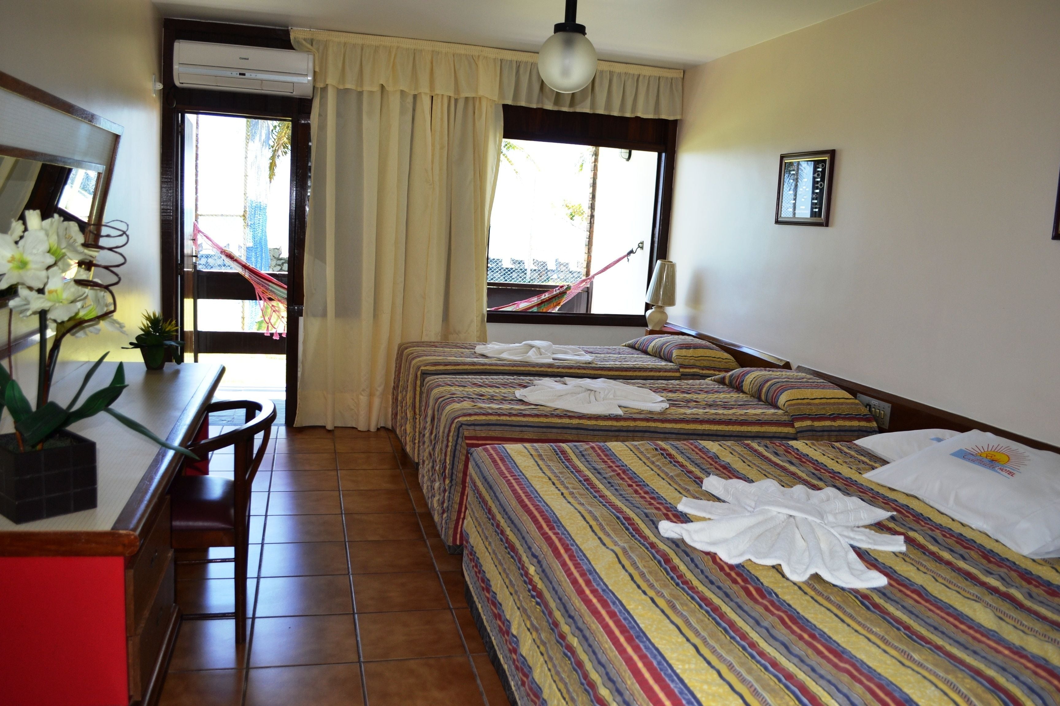 Ein Hotelzimmer mit zwei Betten, einem Schreibtisch mit Stuhl, einem Fenster mit Vorhängen und einer Hängematte im Freien.